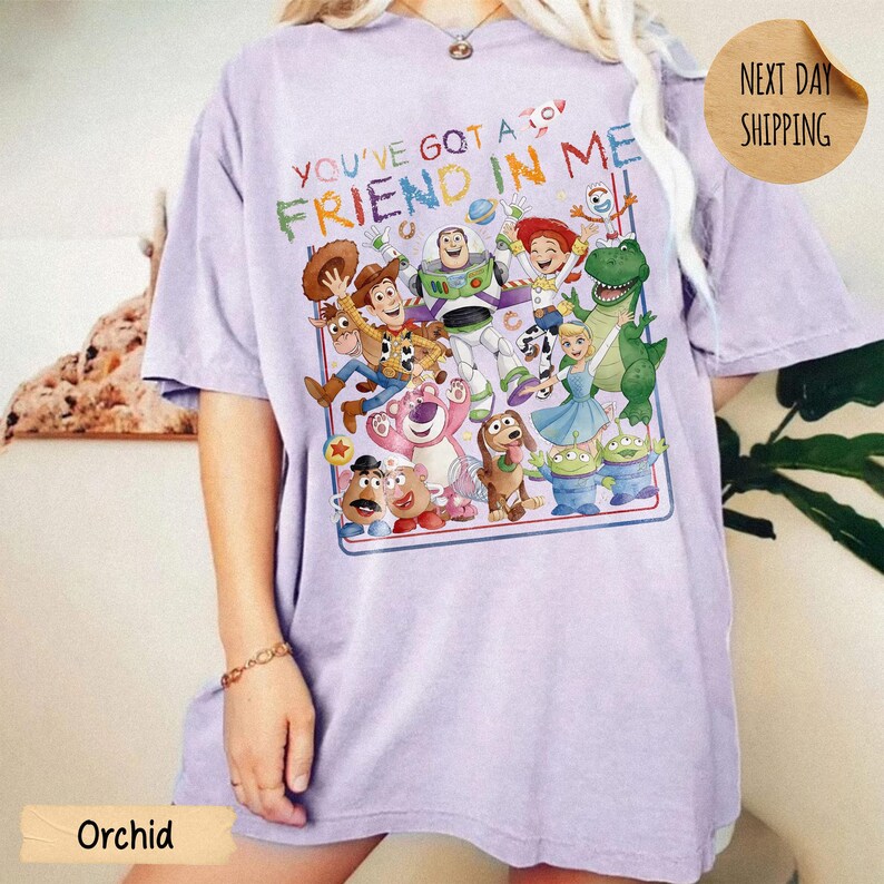 May include: A light purple t-shirt featuring a colorful graphic print of various animated characters from the Toy Story franchise. The text "YOU'VE GOT A FRIEND IN ME" is printed in multi-colored letters above the group of characters.