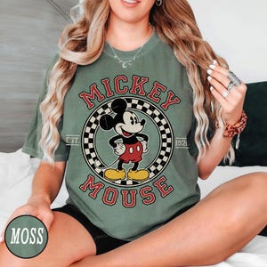 Puede incluir: Camiseta verde musgo con un dise&ntilde;o de Mickey Mouse en un c&iacute;rculo. El dise&ntilde;o incluye las palabras "Mickey Mouse" y "Est. 1928" en rojo y negro. Mickey lleva pantalones cortos rojos y zapatos amarillos.