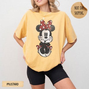 May include: A mustard yellow short-sleeve t-shirt featuring a distressed graphic print of a cartoon mouse character with a red and white polka dot bow. The shirt has a crew neckline and a loose fit. A circular badge on the top right reads NEXT DAY SHIPPING.