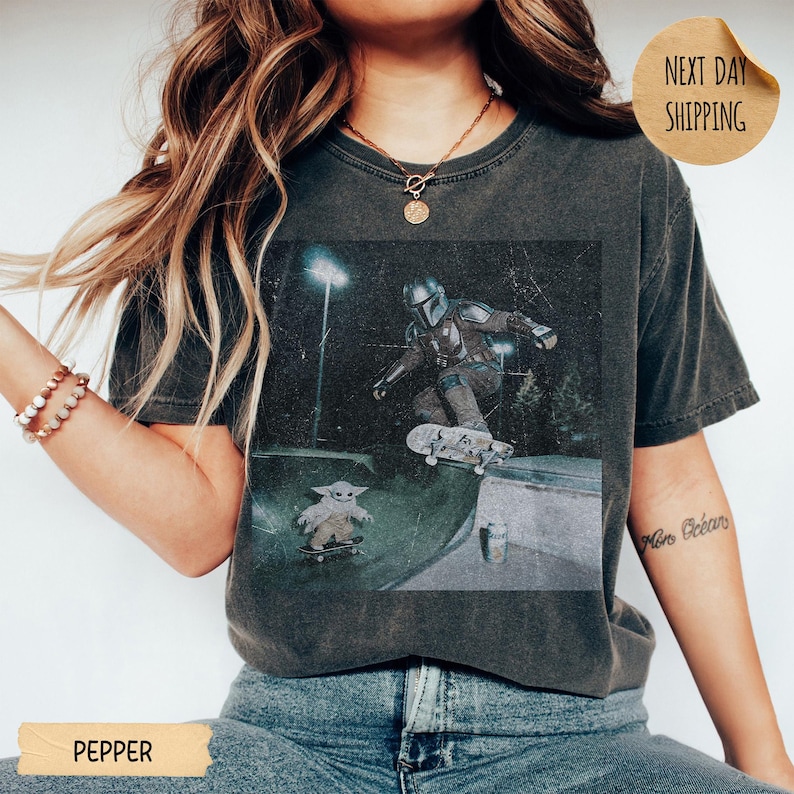 May include: Dark gray t-shirt featuring a graphic of The Mandalorian and Grogu skateboarding in a skate park. The image has a vintage, distressed look. The shirt has a crew neck and short sleeves. The word "PEPPER" is printed at the bottom.