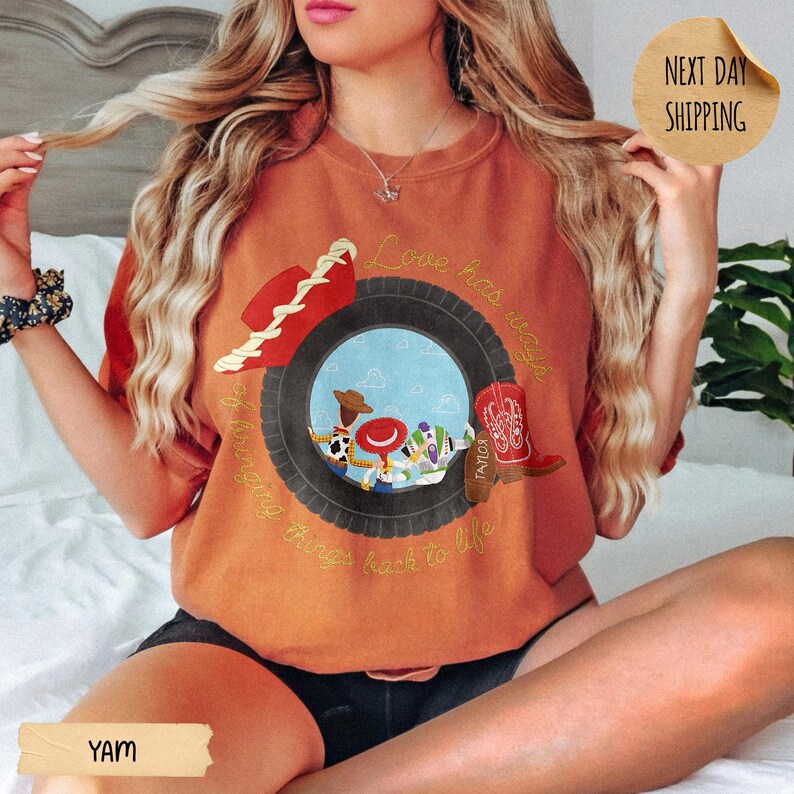 May include: A burnt orange crewneck sweatshirt featuring a graphic of a black tire framing a blue sky scene with cartoon characters, a red cowboy hat, and a red cowboy boot. The text "Love has ways of bringing things back to life" is printed.