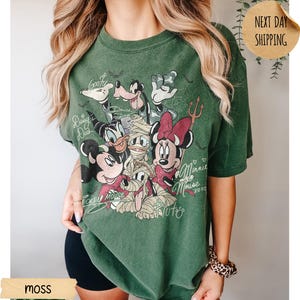 Peut inclure: T-shirt vert mousse avec un motif vintage de personnages Disney, dont Mickey Mouse, Minnie Mouse, Donald Duck, Dingo et Pluto. Le motif comprend les inscriptions "Mickey Mouse", "Minnie Mouse" et "Pluto".