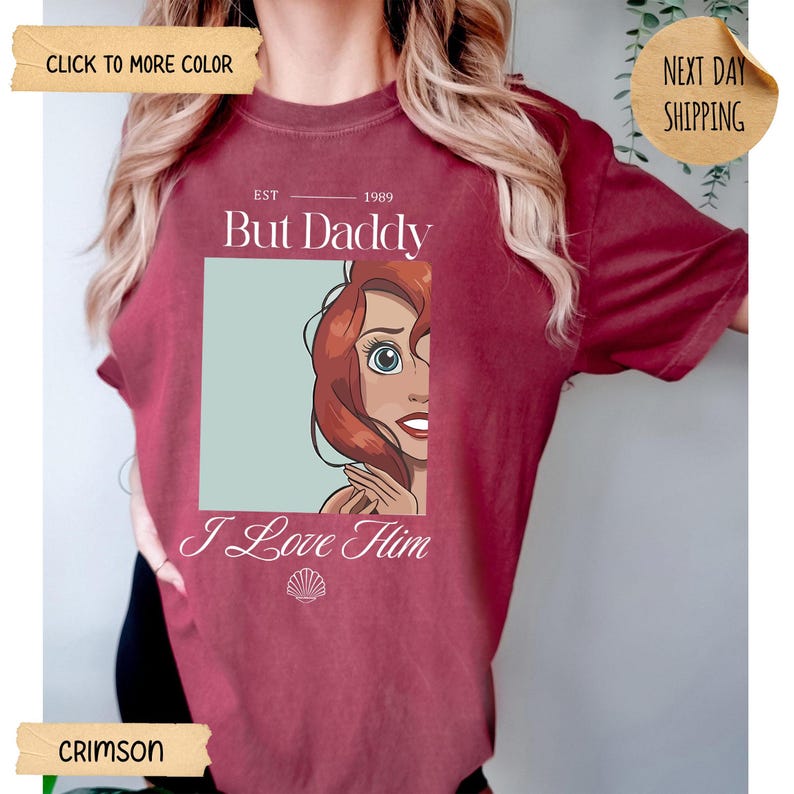 Op de afbeelding: Een karmozijnrood t-shirt met een grafische print van een tekenfilmfiguur met rood haar. De tekst "EST 1989 But Daddy I Love Him" staat boven en onder de afbeelding, met een klein schelppictogram onderaan het grafische ontwerp.