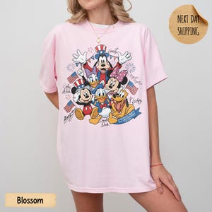 May include: A light pink t-shirt featuring a graphic print of Disney characters including Mickey Mouse, Minnie Mouse, Donald Duck, Daisy Duck, Goofy, and Pluto wearing patriotic attire, with American flags and the text America 250th Anniversary.