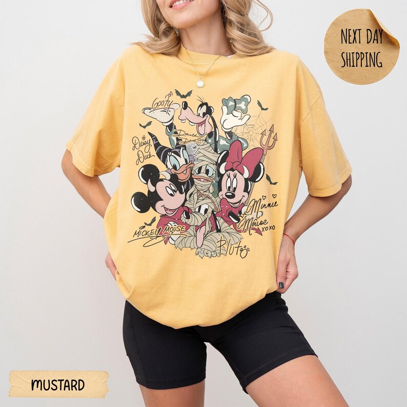 Peut inclure: T-shirt jaune moutarde avec un motif de personnages Disney d&eacute;guis&eacute;s en momies. Le motif comprend Mickey Mouse, Minnie Mouse, Donald Duck, Dingo et Pluto, avec leurs noms et signatures. Le t-shirt porte &eacute;galement l'inscription "Next Day Shipping".