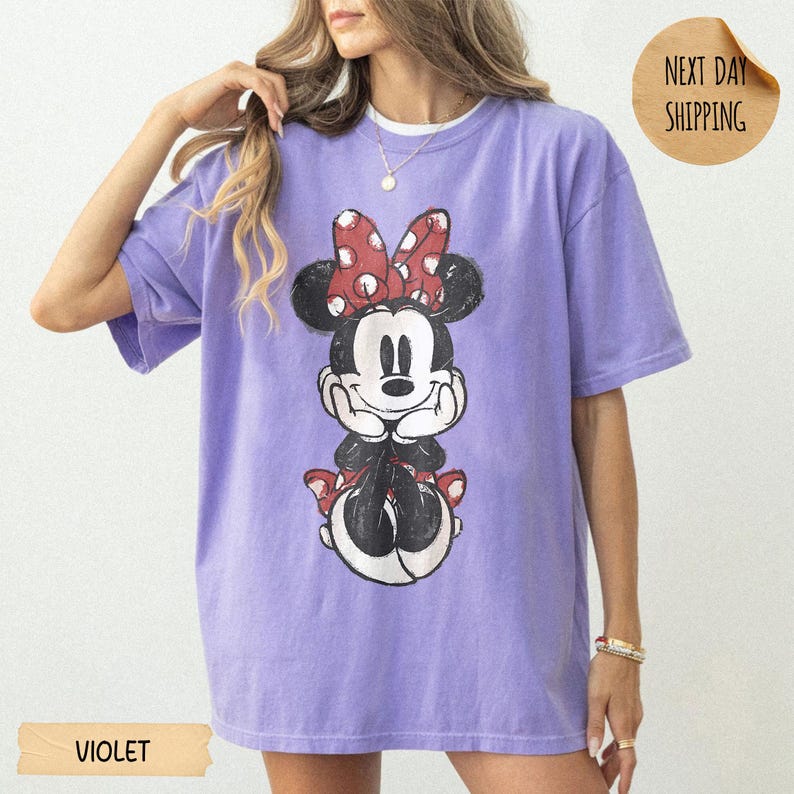 May include: A violet short-sleeved t-shirt featuring a distressed graphic print of a cartoon mouse character with a red and white polka dot bow. The character is depicted with hands resting under the chin, wearing a red skirt and black shoes.