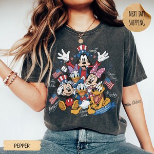 May include: A charcoal grey short-sleeve t-shirt featuring a graphic of Mickey Mouse, Minnie Mouse, Donald Duck, Daisy Duck, Goofy, and Pluto wearing patriotic hats. The design includes American flags and the text America 250 Anniversary 1776-2026.