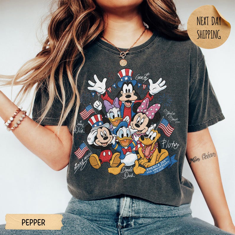 May include: A charcoal grey short-sleeve t-shirt featuring a graphic of Mickey Mouse, Minnie Mouse, Donald Duck, Daisy Duck, Goofy, and Pluto wearing patriotic hats. The design includes American flags and the text America 250 Anniversary 1776-2026.