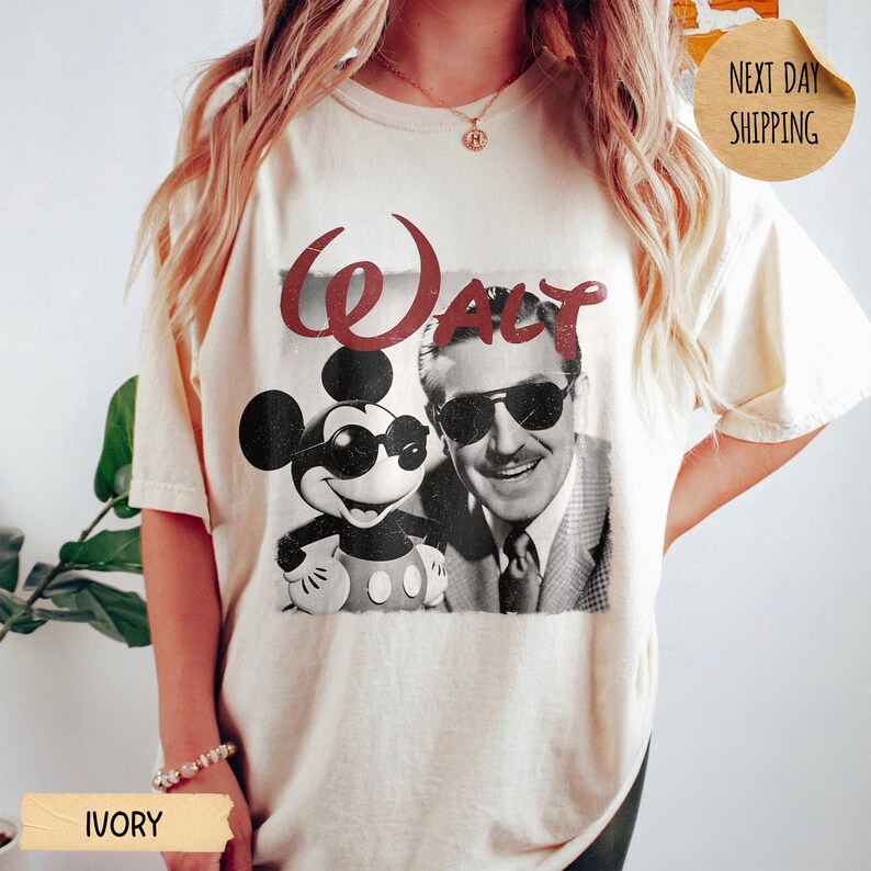 May include: Ivory-colored t-shirt featuring a graphic of Mickey Mouse and Walt Disney, with the word "Walt" in red. The shirt has a classic, vintage design, perfect for Disney fans.