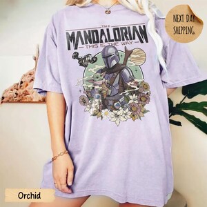 May include: Orchid-colored t-shirt featuring a graphic of The Mandalorian and Grogu surrounded by flowers. The text "THE MANDALORIAN - THIS IS THE WAY" is printed across the top. The shirt has a relaxed fit.