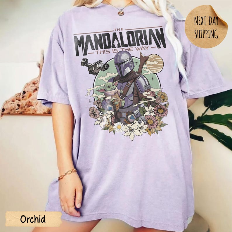May include: Orchid-colored t-shirt featuring a graphic of The Mandalorian and Grogu surrounded by flowers. The text "THE MANDALORIAN - THIS IS THE WAY" is printed across the top. The shirt has a relaxed fit.
