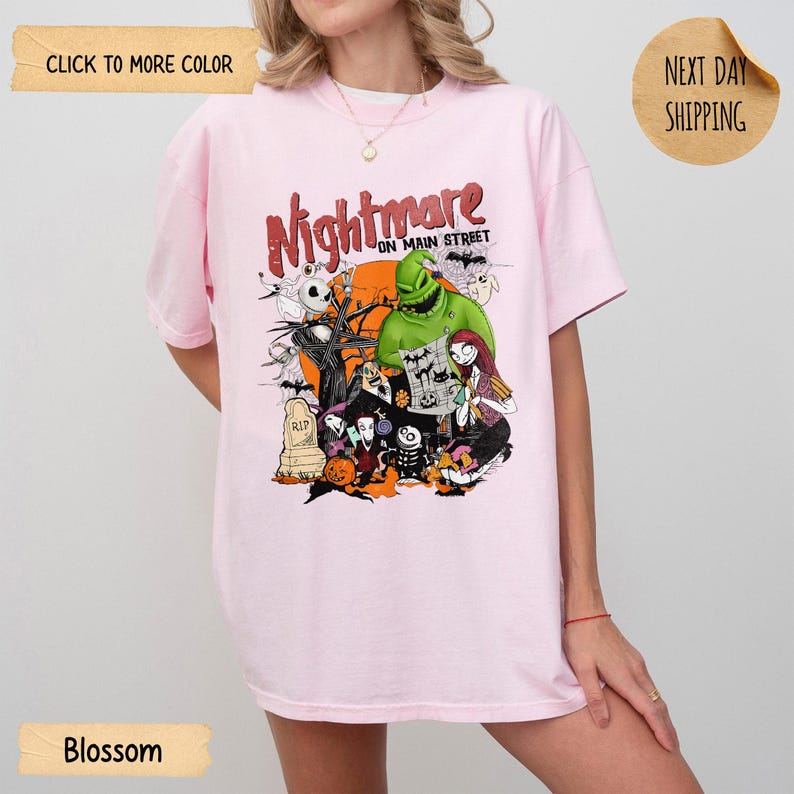 May include: A light pink t-shirt featuring a graphic print with the text "Nightmare on Main Street" and various characters including a green creature, a skeleton, and a female figure, set against an orange moon and dark tree silhouettes.