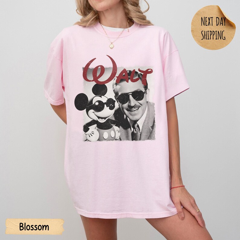 May include: Light pink t-shirt featuring a black and white graphic of Mickey Mouse and Walt Disney with the word "Walt" in red. The shirt has short sleeves and a relaxed fit. The word "Blossom" is printed on a light yellow rectangle.