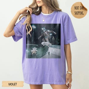 May include: Violet-colored t-shirt featuring a graphic of a character skateboarding, with another character on a skateboard below. The image has a distressed, vintage look. The shirt has a relaxed fit and short sleeves. The word "VIOLET" is printed at the bottom.