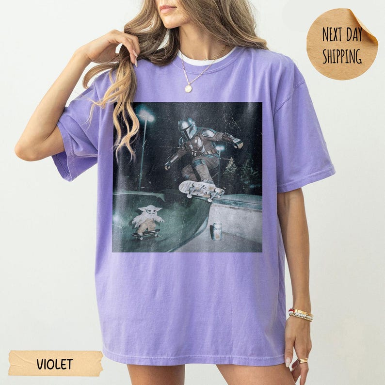 May include: Violet-colored t-shirt featuring a graphic of a character skateboarding, with another character on a skateboard below. The image has a distressed, vintage look. The shirt has a relaxed fit and short sleeves. The word "VIOLET" is printed at the bottom.