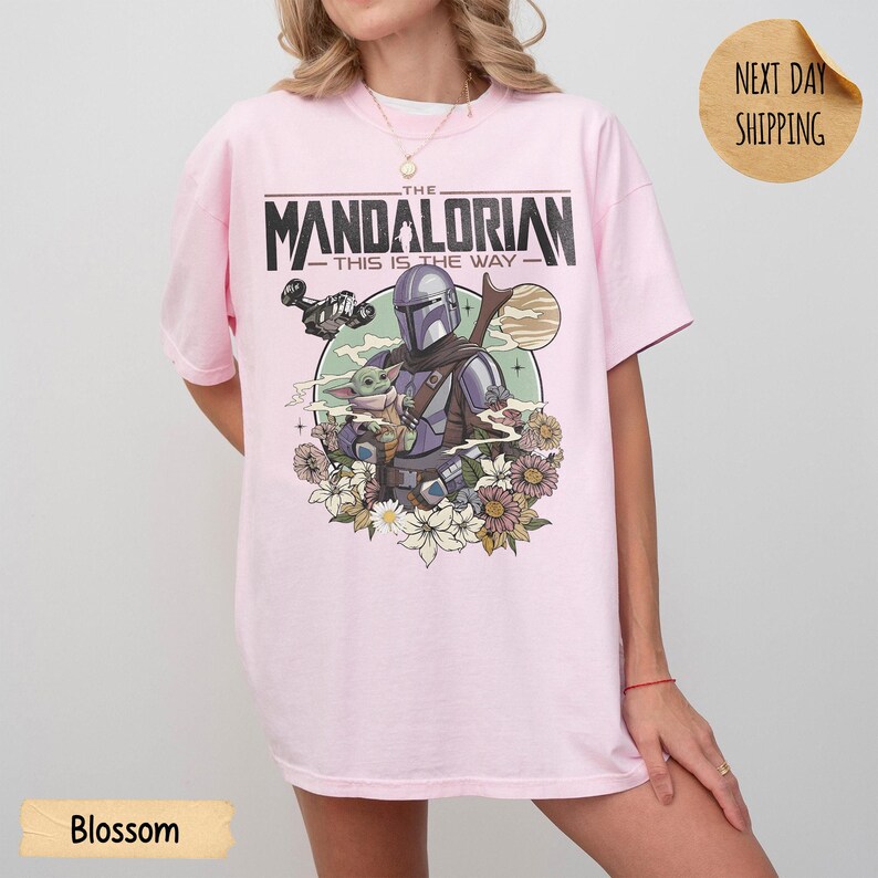 May include: A light pink t-shirt featuring a graphic of The Mandalorian and Grogu surrounded by flowers. The text "THE MANDALORIAN - THIS IS THE WAY" is printed across the top. The shirt is a relaxed fit.