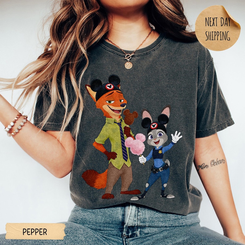 May include: Dark gray t-shirt featuring a cartoon illustration of Nick Wilde and Judy Hopps from Zootopia, each wearing Mickey Mouse ears. Nick holds a chocolate treat, and Judy holds a pink cotton candy. The word "PEPPER" is at the bottom.