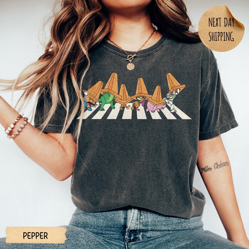 May include: A charcoal grey t-shirt featuring a graphic of five animated characters walking across a white striped crosswalk, each wearing an orange traffic cone on their head. The design includes Mr. Potato Head, Rex, Hamm, and Buzz Lightyear.