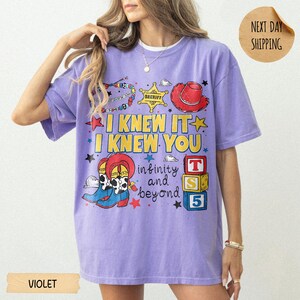 May include: A violet short-sleeve t-shirt featuring a graphic print with the text I KNEW IT I KNEW YOU infinity and beyond. The design includes a sheriff badge, a red cowboy hat, cowboy boots, and three stacked blocks labeled T, S, and 5.
