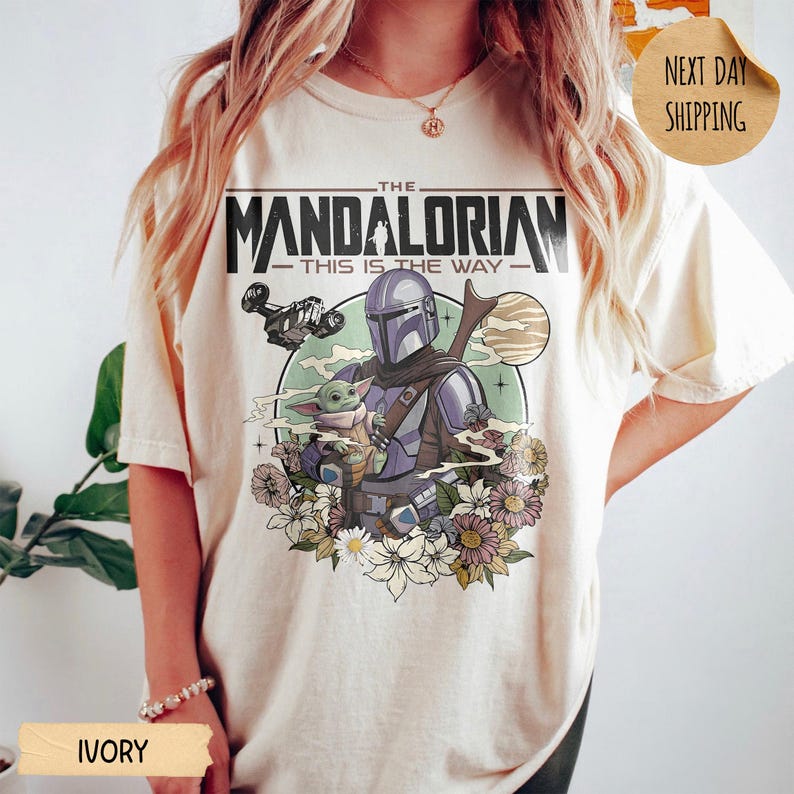 May include: Ivory-colored t-shirt featuring a graphic of The Mandalorian and Grogu, surrounded by flowers. The design includes the text "THE MANDALORIAN - THIS IS THE WAY" and a spaceship. The shirt has a crew neck and short sleeves.