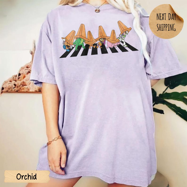 May include: A light purple short-sleeve t-shirt featuring a graphic print of five cartoon characters walking across a black and white striped zebra crossing, each wearing an orange traffic cone on their head as a hat.