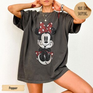 May include: A charcoal gray oversized t-shirt featuring a distressed graphic print of Minnie Mouse with a red and white polka dot bow. The character is depicted with hands resting under the chin. The shirt has short sleeves and a crew neckline.