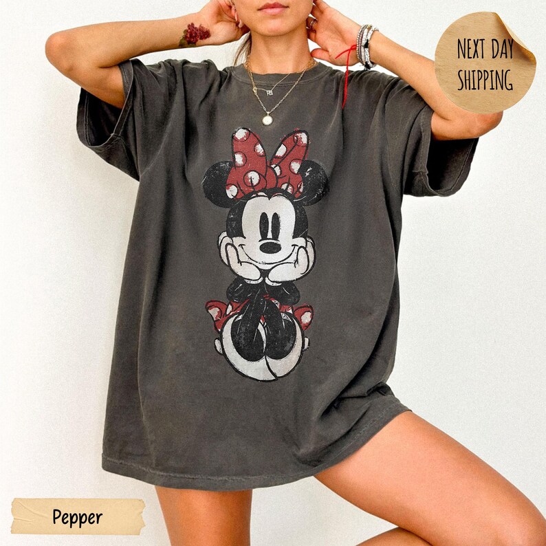 May include: A charcoal gray oversized t-shirt featuring a distressed graphic print of Minnie Mouse with a red and white polka dot bow. The character is depicted with hands resting under the chin. The shirt has short sleeves and a crew neckline.