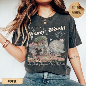 May include: Dark gray t-shirt with a vintage-style graphic of Disney World in Florida. The design includes Mickey Mouse, a castle, Spaceship Earth, and the text "Florida Disney World Est. 1971". The shirt also has the text "The Most Magical Place On Earth".