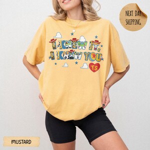 May include: A mustard yellow short-sleeve t-shirt featuring a graphic print with the text I KNEW IT, I KNEW YOU. The design includes cartoon character patterns, small blue stars, white clouds, and a red heart with the letters TS inside.