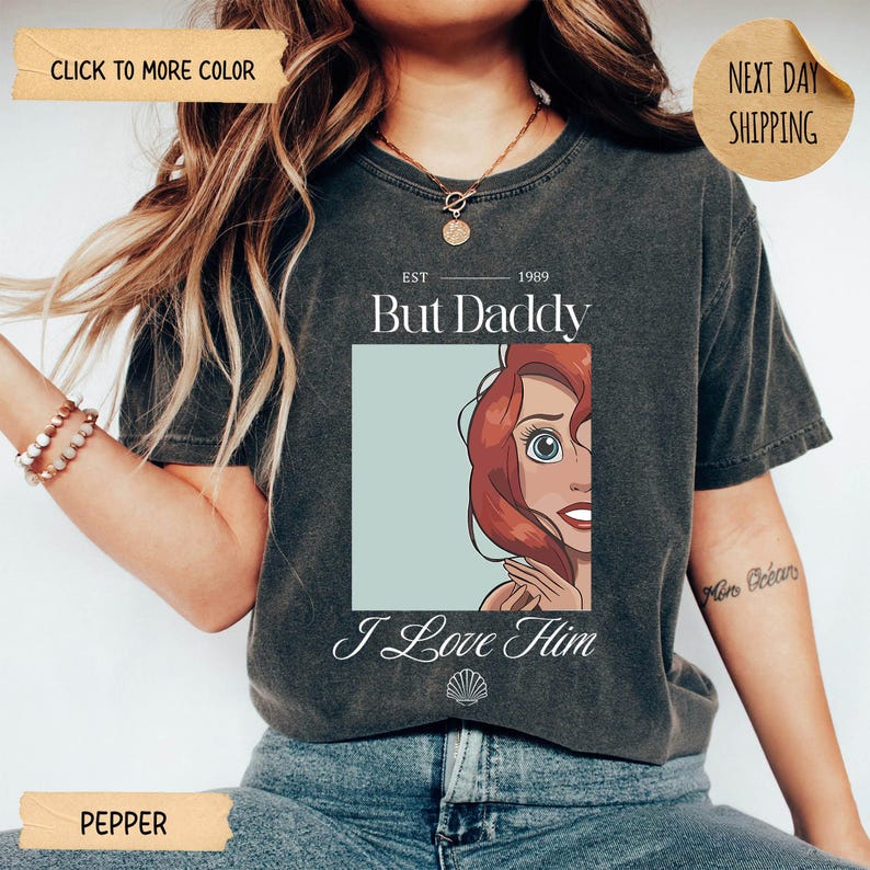 Op de afbeelding: Een antracietgrijs T-shirt met korte mouwen en een grafische print van een roodharig personage in een lichtblauw vierkant. De tekst luidt EST 1989 But Daddy I Love Him, met een klein schelppictogram onder het grafische ontwerp.