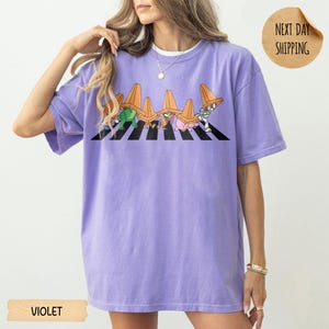 May include: A light purple short-sleeve t-shirt featuring a graphic print of cartoon characters walking across a black and white striped zebra crossing, each wearing an orange traffic cone on their head. The shirt has a crew neckline and a loose fit.