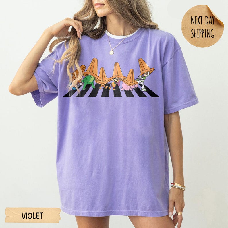 May include: A light purple short-sleeve t-shirt featuring a graphic print of cartoon characters walking across a black and white striped zebra crossing, each wearing an orange traffic cone on their head. The shirt has a crew neckline and a loose fit.