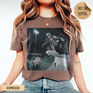 May include: Espresso-colored t-shirt featuring a graphic of The Mandalorian skateboarding, with Grogu on a skateboard below. The image is set in a skate park at night. The shirt has a vintage, worn look. The words "Next Day Shipping" are on a circular sticker.
