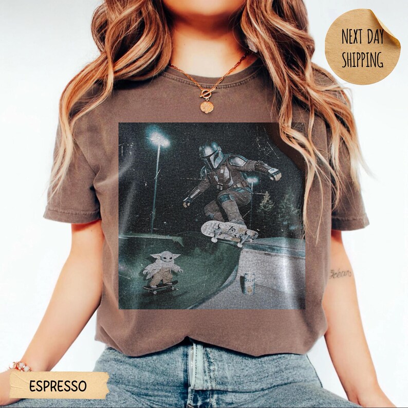 May include: Espresso-colored t-shirt featuring a graphic of The Mandalorian skateboarding, with Grogu on a skateboard below. The image is set in a skate park at night. The shirt has a vintage, worn look. The words "Next Day Shipping" are on a circular sticker.