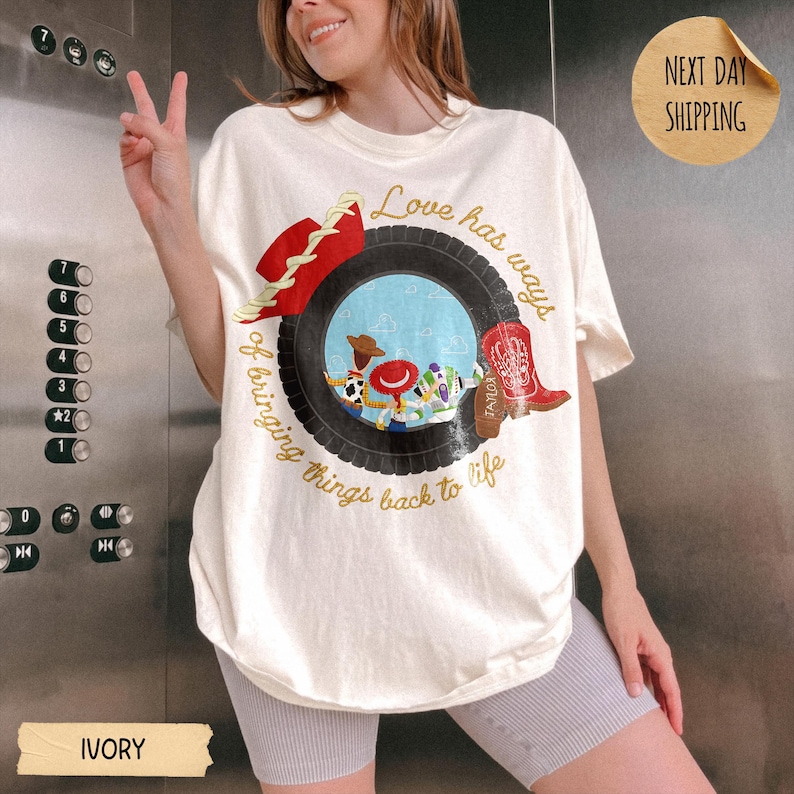 May include: An ivory t-shirt featuring a graphic of a black tire framing a scene with cartoon characters, a red cowboy hat, and a red cowboy boot. The text reads, Love has ways of bringing things back to life, in a cursive gold font.