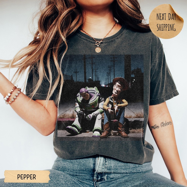 Pode incluir: T-shirt cinza escuro com um gr&aacute;fico de Buzz Lightyear e Woody de Toy Story sentados em uma cal&ccedil;ada &agrave; noite. A camisa tem um visual vintage e desgastado. A palavra "PEPPER" est&aacute; impressa na parte inferior.