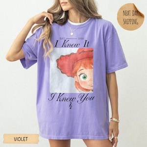 May include: A violet short-sleeved t-shirt featuring a graphic print of a cartoon character with red hair and a red hat. The text reads "EST 2026 I Knew It" above the graphic and "I Knew You" below it, with a small musical note symbol included.