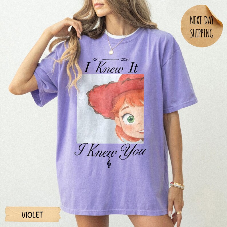 May include: A violet short-sleeved t-shirt featuring a graphic print of a cartoon character with red hair and a red hat. The text reads "EST 2026 I Knew It" above the graphic and "I Knew You" below it, with a small musical note symbol included.