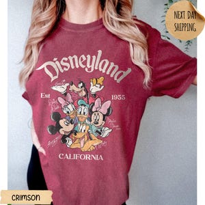 May include: A crimson short-sleeve t-shirt featuring a graphic print of Disneyland, Est 1955, California. The design includes illustrations of Mickey Mouse, Minnie Mouse, Donald Duck, Daisy Duck, Goofy, and Pluto with their names written in script.