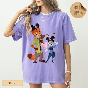 May include: Violet-colored t-shirt featuring a cartoon illustration of Nick Wilde and Judy Hopps from Zootopia. Nick is wearing a green shirt and Judy is in a police uniform. The shirt has the word "VIOLET" printed on the bottom.