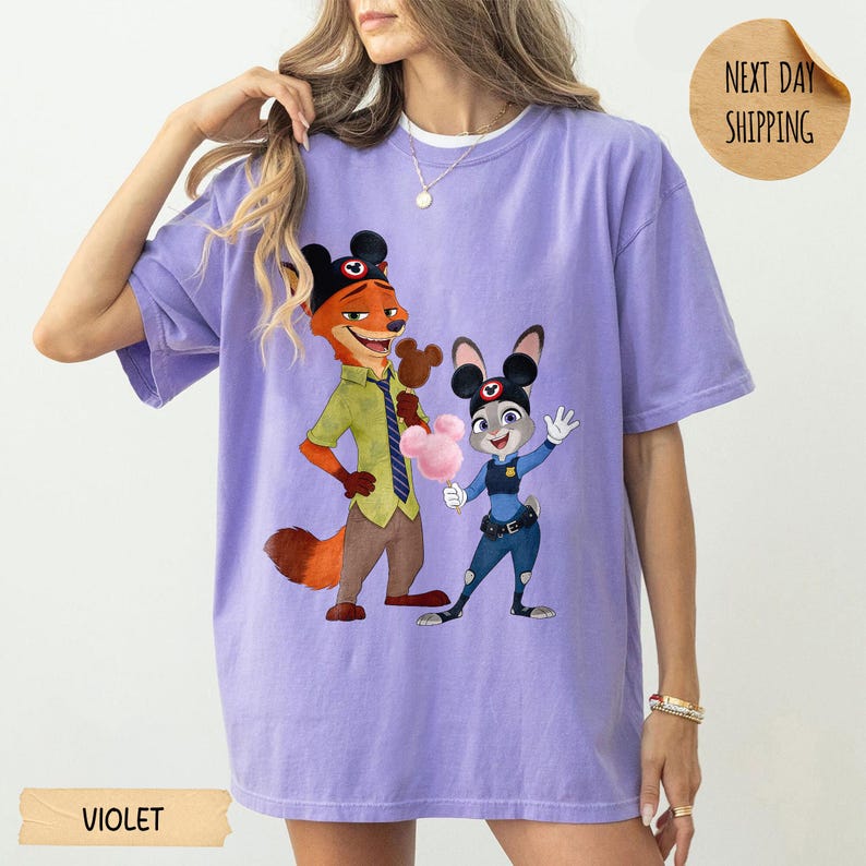 May include: Violet-colored t-shirt featuring a cartoon illustration of Nick Wilde and Judy Hopps from Zootopia. Nick is wearing a green shirt and Judy is in a police uniform. The shirt has the word "VIOLET" printed on the bottom.