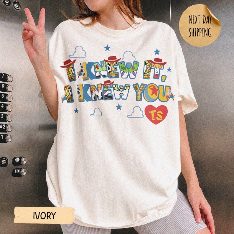 May include: An ivory short-sleeve t-shirt featuring a graphic print with the text I KNEW IT, I KNEW YOU and TS inside a red heart. The letters are decorated with patterns inspired by toy characters, including cowboy hats, space ranger suits, and stars.