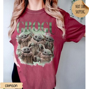 May include: A crimson short-sleeve t-shirt featuring a graphic print of five small green creatures in tan robes. The text GROGU is printed in large, distressed green letters above the characters, who are depicted in various poses.