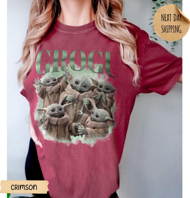May include: A crimson short-sleeve t-shirt featuring a graphic print of five small green creatures in tan robes. The text GROGU is printed in large, distressed green letters above the characters, who are depicted in various poses.