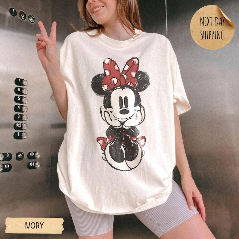 May include: An ivory short-sleeve t-shirt featuring a distressed graphic print of Minnie Mouse wearing a red and white polka dot bow. The character is depicted with hands resting under the chin. The shirt has a crew neckline and a loose fit.