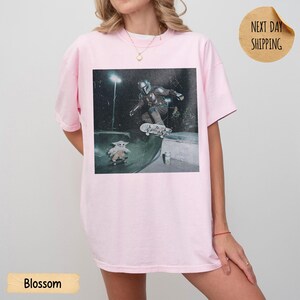 May include: Light pink t-shirt with a graphic of a character from The Mandalorian skateboarding in a skate park. The shirt has short sleeves and a crew neck. The word "Blossom" is printed at the bottom of the shirt.