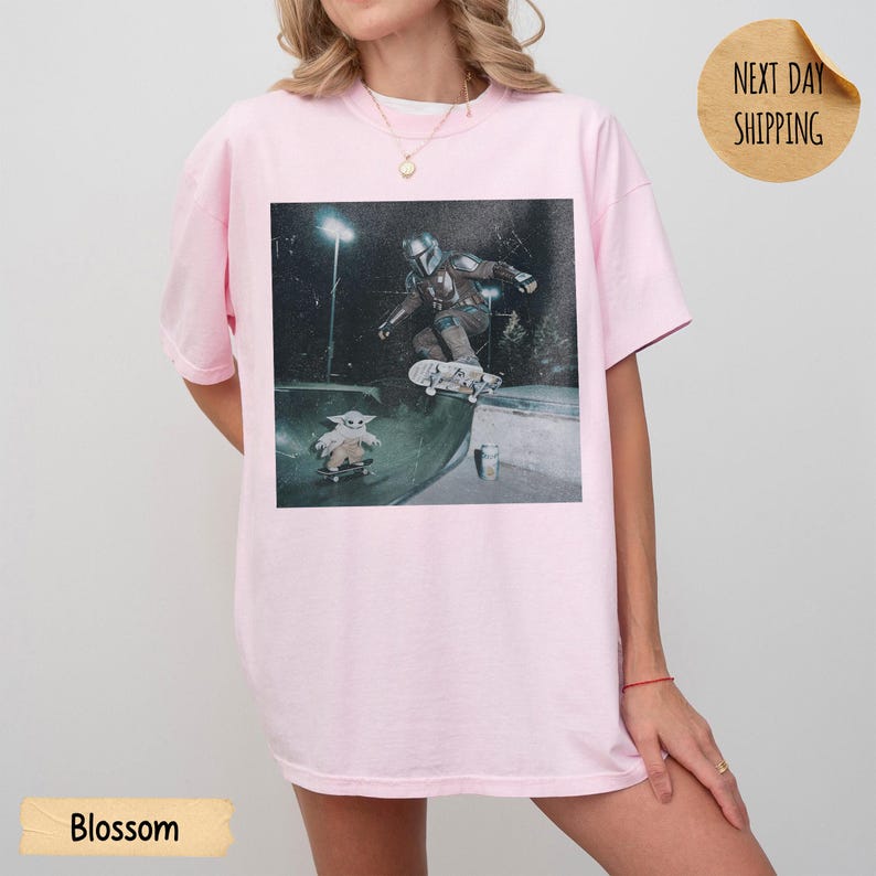 May include: Light pink t-shirt with a graphic of a character from The Mandalorian skateboarding in a skate park. The shirt has short sleeves and a crew neck. The word "Blossom" is printed at the bottom of the shirt.