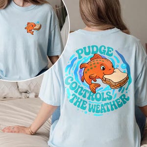 May include: Pale blue t-shirt with a cartoon orange fish and sandwich graphic. The back of the shirt has the text "Pudge Controls the Weather" in blue and green. A smaller version of the fish and sandwich graphic is on the front.
