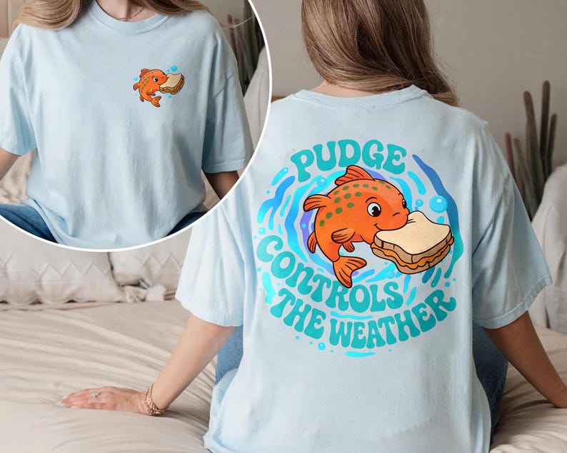 May include: Pale blue t-shirt with a cartoon orange fish and sandwich graphic. The back of the shirt has the text "Pudge Controls the Weather" in blue and green. A smaller version of the fish and sandwich graphic is on the front.