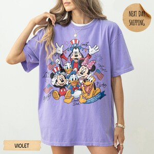 May include: A violet short-sleeve t-shirt featuring a graphic print of Mickey Mouse, Minnie Mouse, Donald Duck, Daisy Duck, Goofy, and Pluto wearing patriotic hats. The design includes American flags, fireworks, and text reading America 250th.